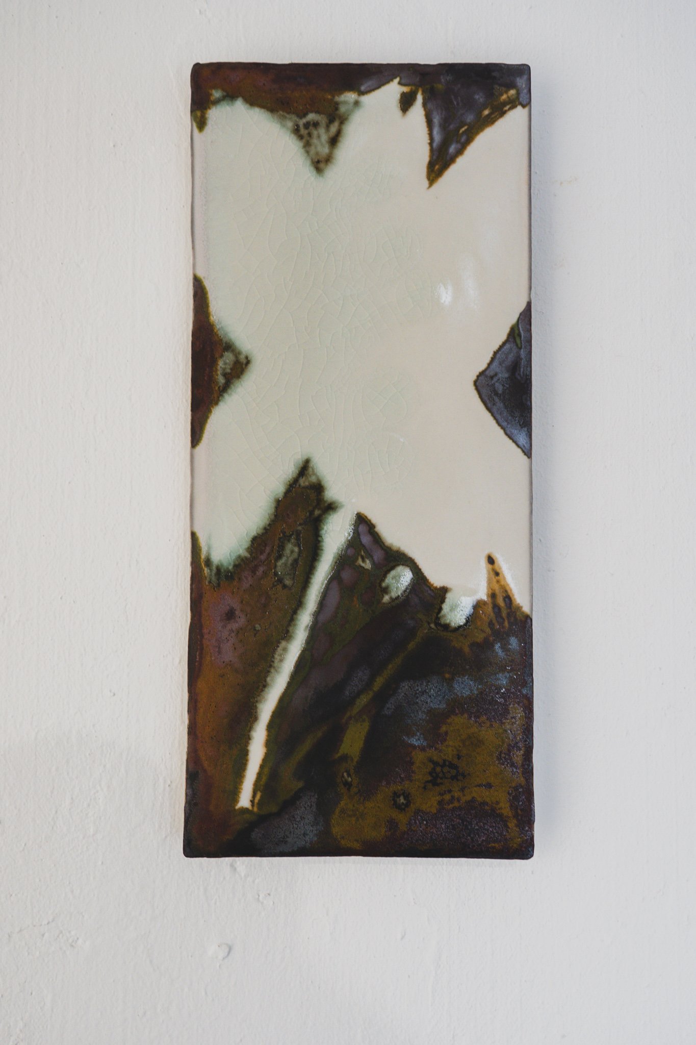 Narrow rectangular ceramic wall panel with an abstract iron-oxide glaze composition, rust-brown and purple bleeding across a pale cream background