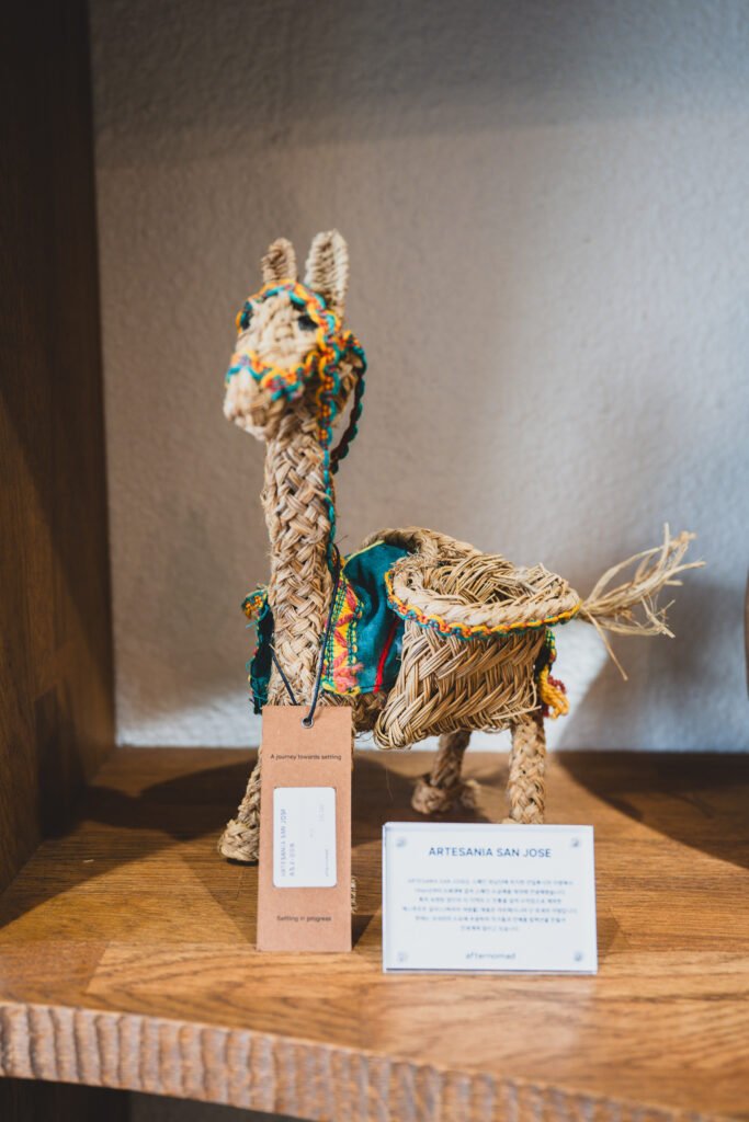 Woven straw llama figure with an "afternomad" tagline card reading "A journey towards settling / Settling in progress," next to an Artesania San Jose product card