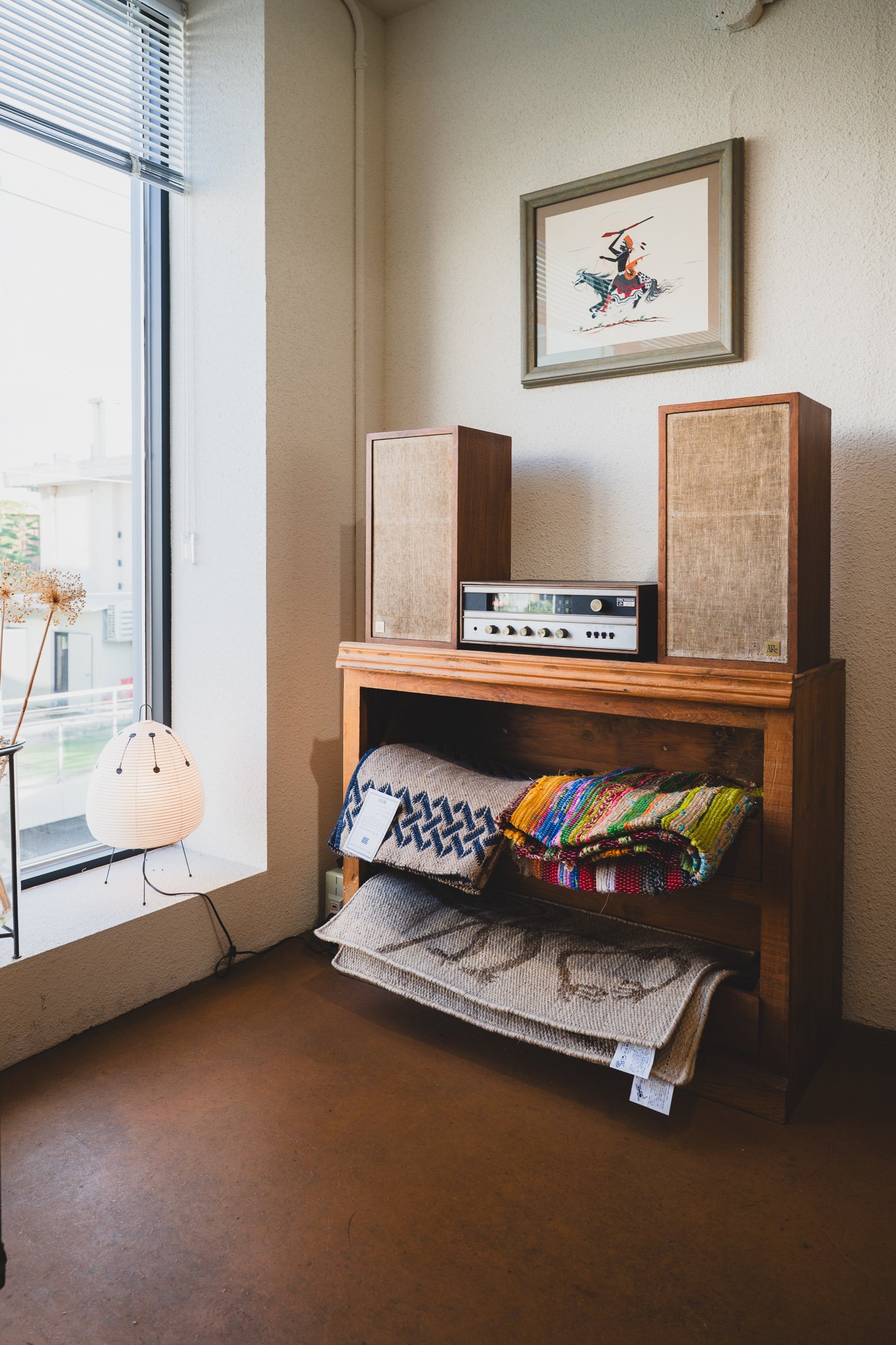 Vintage speakers and a receiver on a wooden cabinet, a framed illustration of a mounted African warrior above, colorful African-pattern woven rugs and fabric folded below