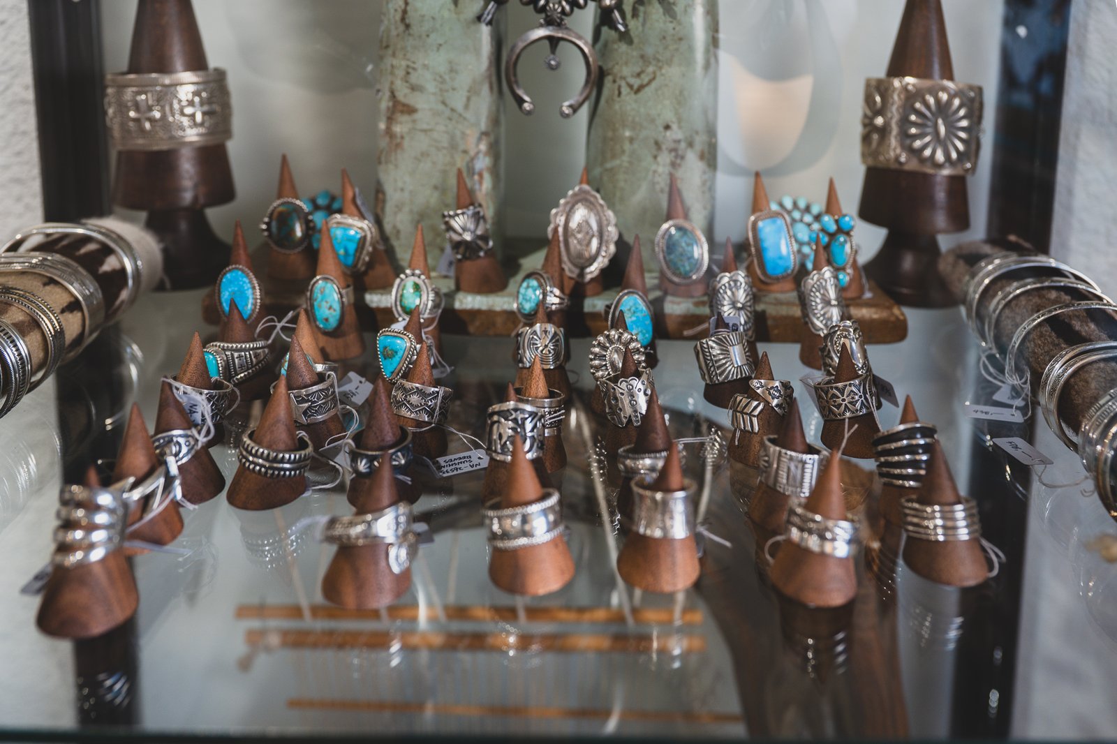 Turquoise and silver rings and cuffs displayed on wooden cone stands inside a glass case, with a squash-blossom-style necklace visible in the background