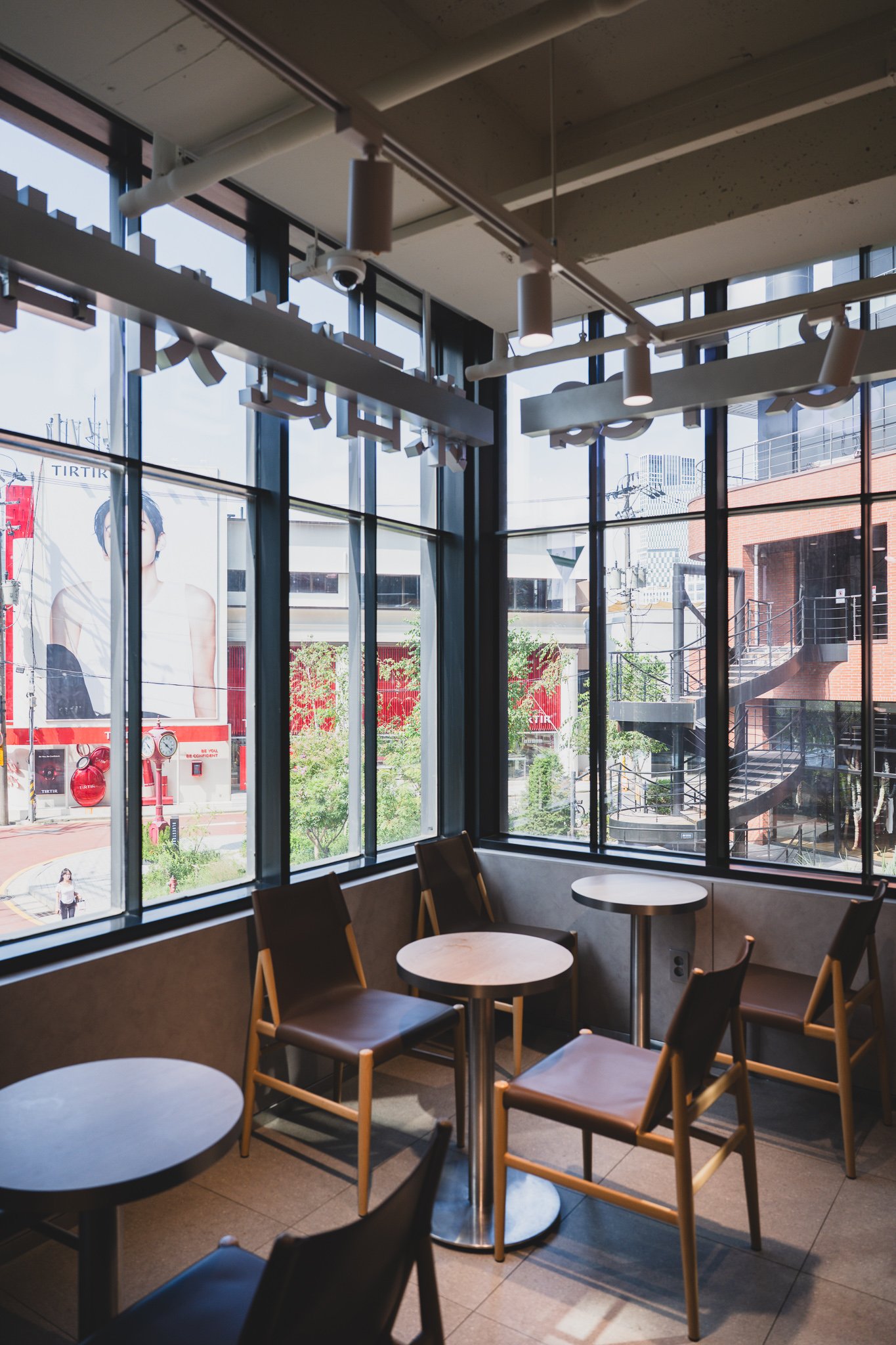 View from a window corner seat overlooking the Seongsu street below, with a TIRTIR billboard and a spiral staircase on a brick building across the street