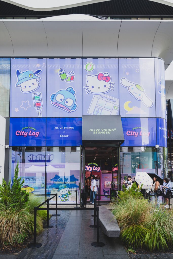 Storefront of Olive Young N Seongsu wrapped in purple City Log signage featuring Hello Kitty, Cinnamoroll, and Hangyodon, with a short line of visitors waiting at the entrance under a "City Log" awning
