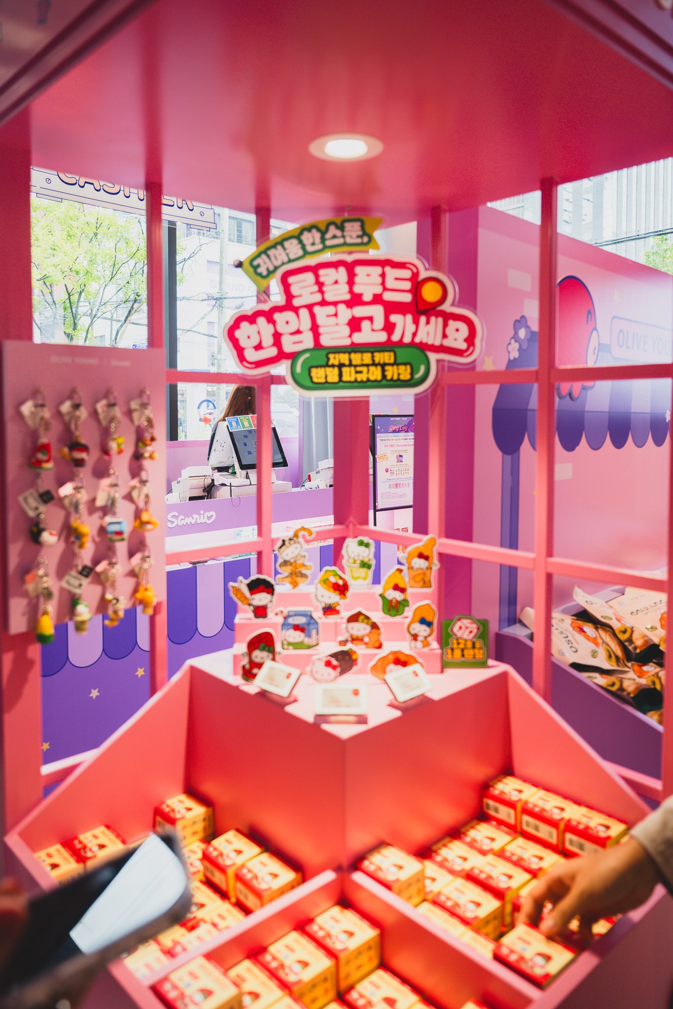 Gacha station signage advertising a random local-themed Hello Kitty figure keyring giveaway, with prize boxes stacked below