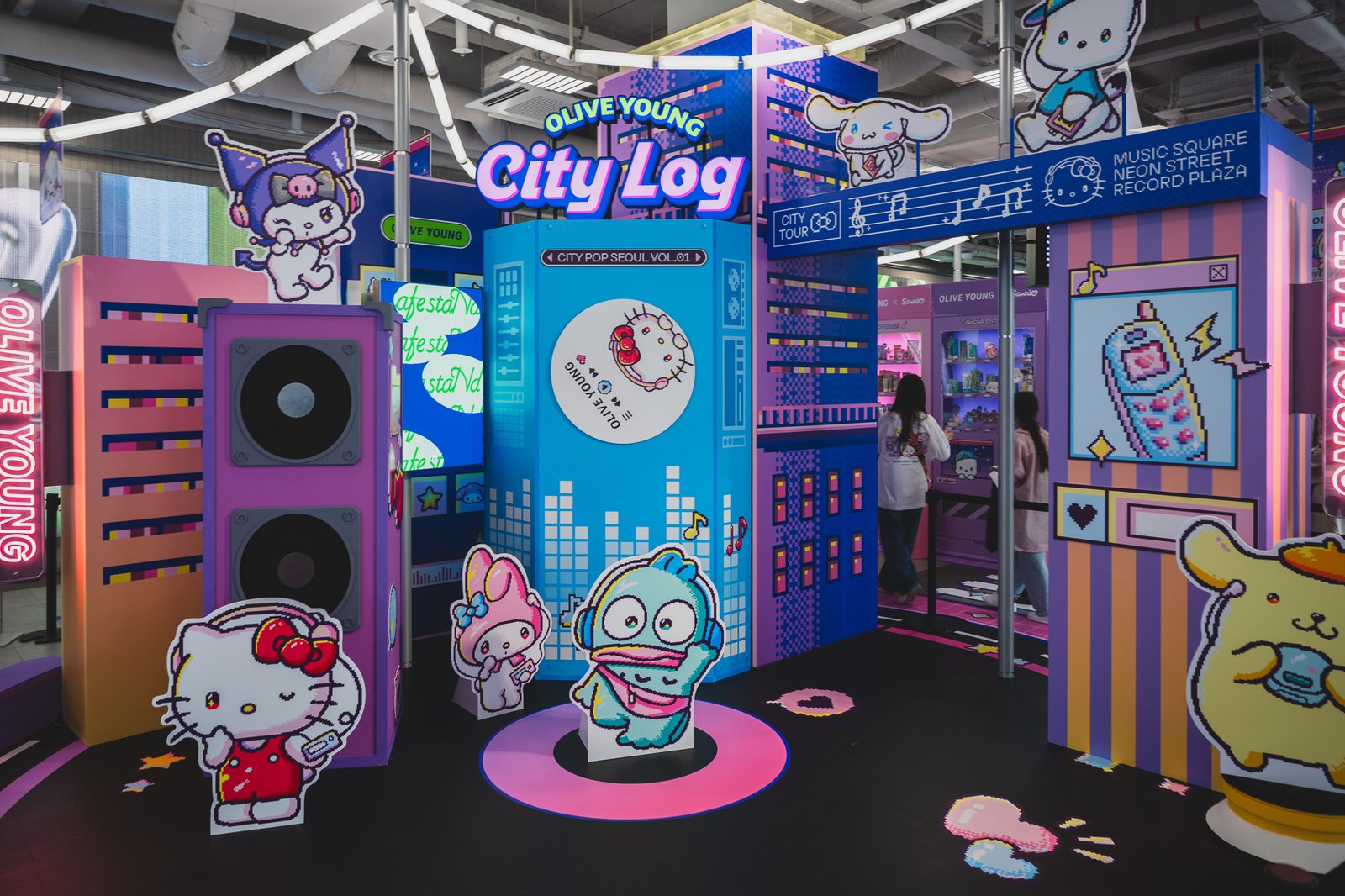 City Log experience zone with pixel-art skyline graphics, an oversized speaker prop, and a record-shaped display, with Hello Kitty and My Melody character cutouts wearing headphones