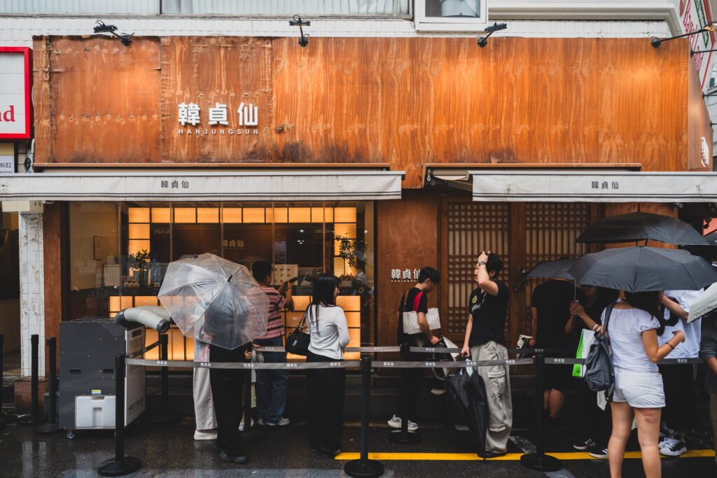 Hanjungsun's Seongsu storefront with a corten-steel and wood exterior sign, and a line of customers with umbrellas waiting outside on a rainy day