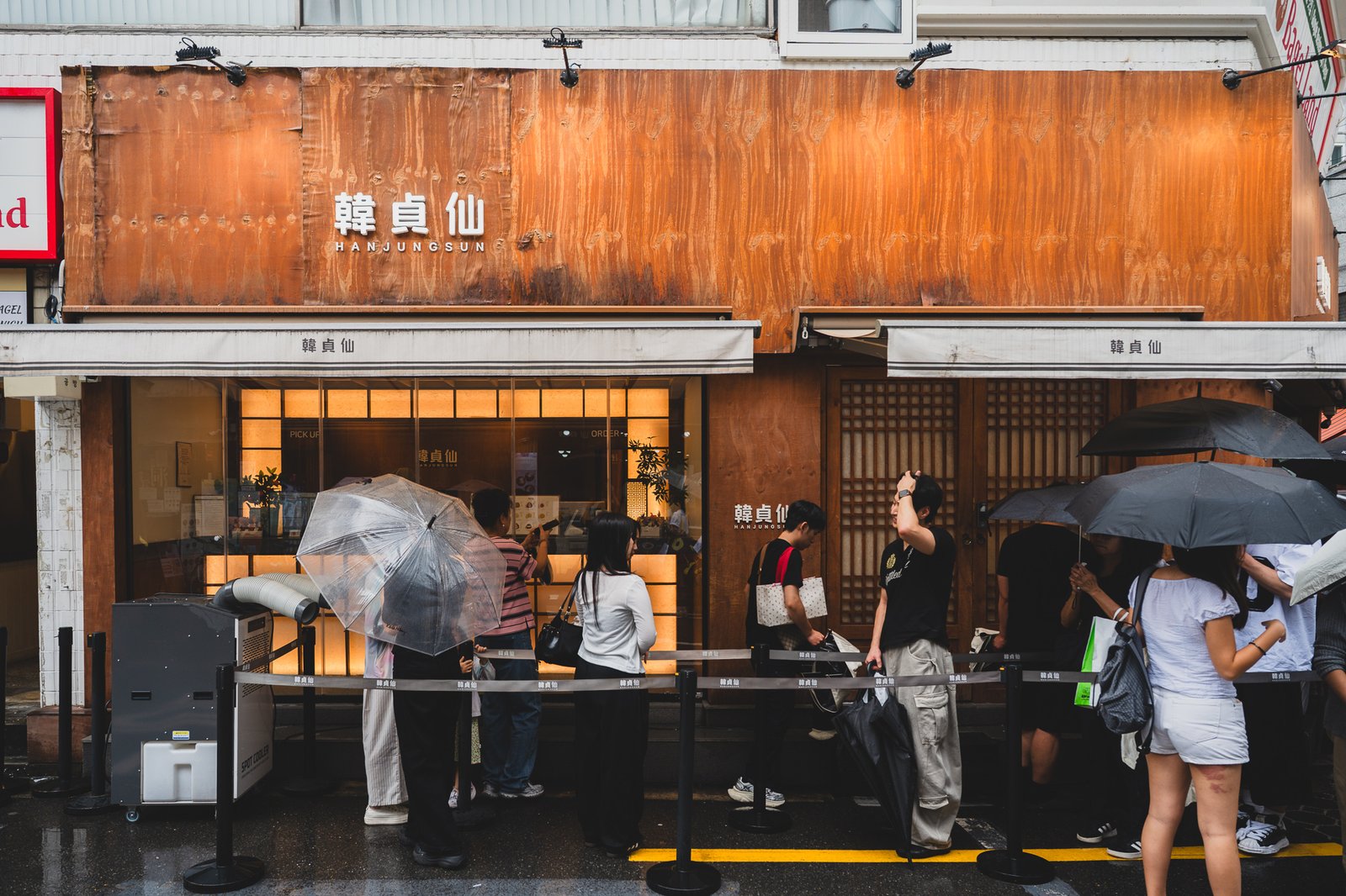 Hanjungsun's Seongsu storefront with a corten-steel and wood exterior sign, and a line of customers with umbrellas waiting outside on a rainy day