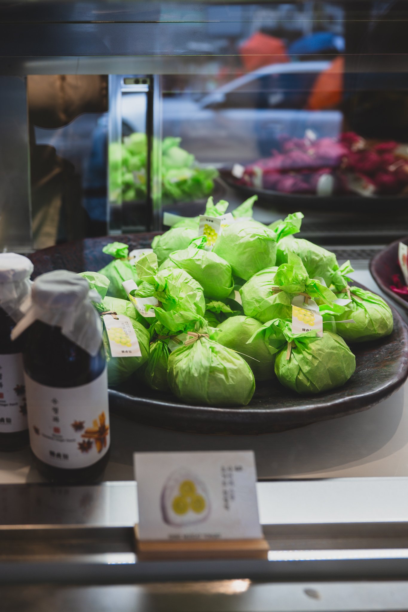 Rows of individually wrapped mochi in green, magenta, and gold paper, arranged on a wooden tray