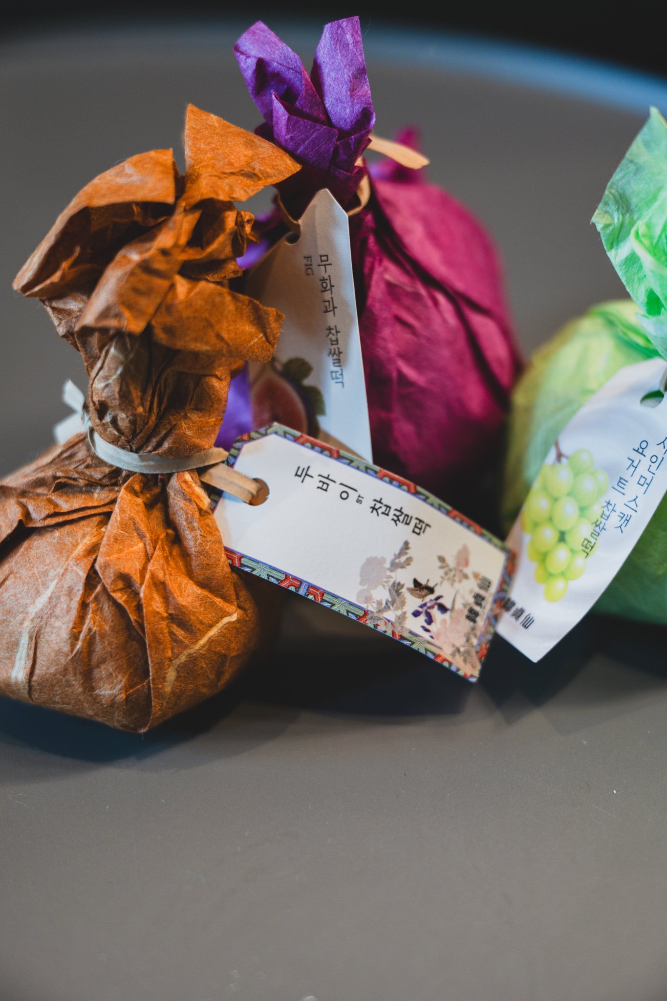 Three mochi still in their colored paper wrapping — brown, magenta, and green — on a table