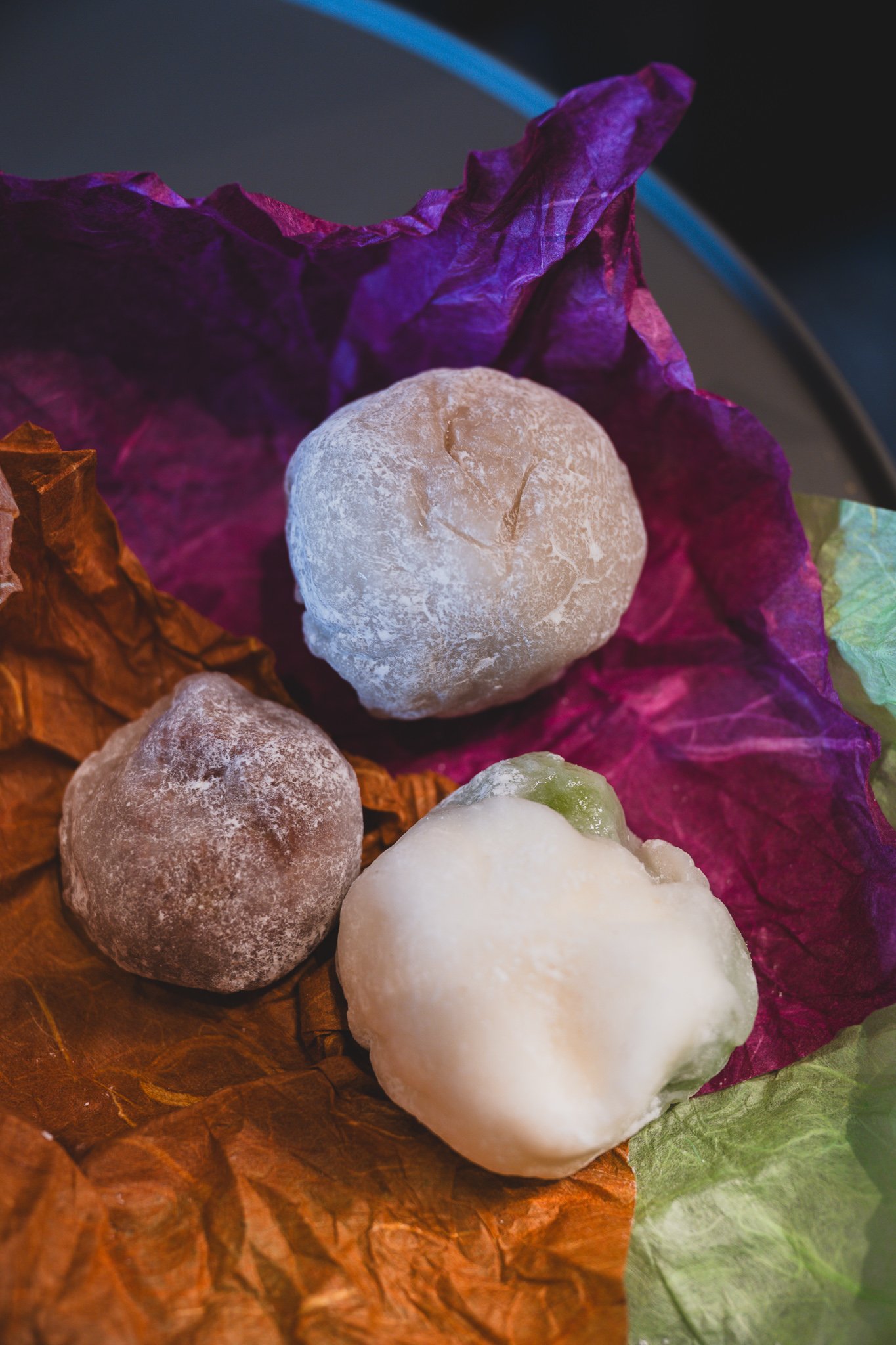 Three unwrapped mochi sitting on their colored wrapping paper — a pale beige one, a darker dusted one, and a plain white one