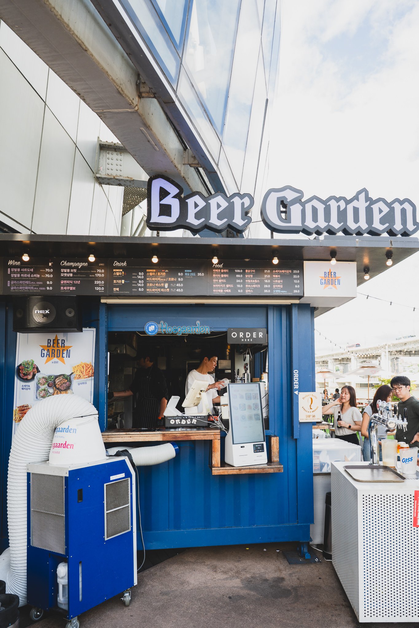 Blue container-style order booth with a Hoegaarden neon sign and a full menu board