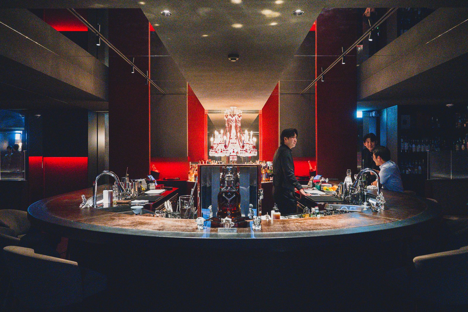 The round bar counter at Baccarat Bar Seoul, wrapped in red lighting and crystal chandeliers, with a bartender working behind it