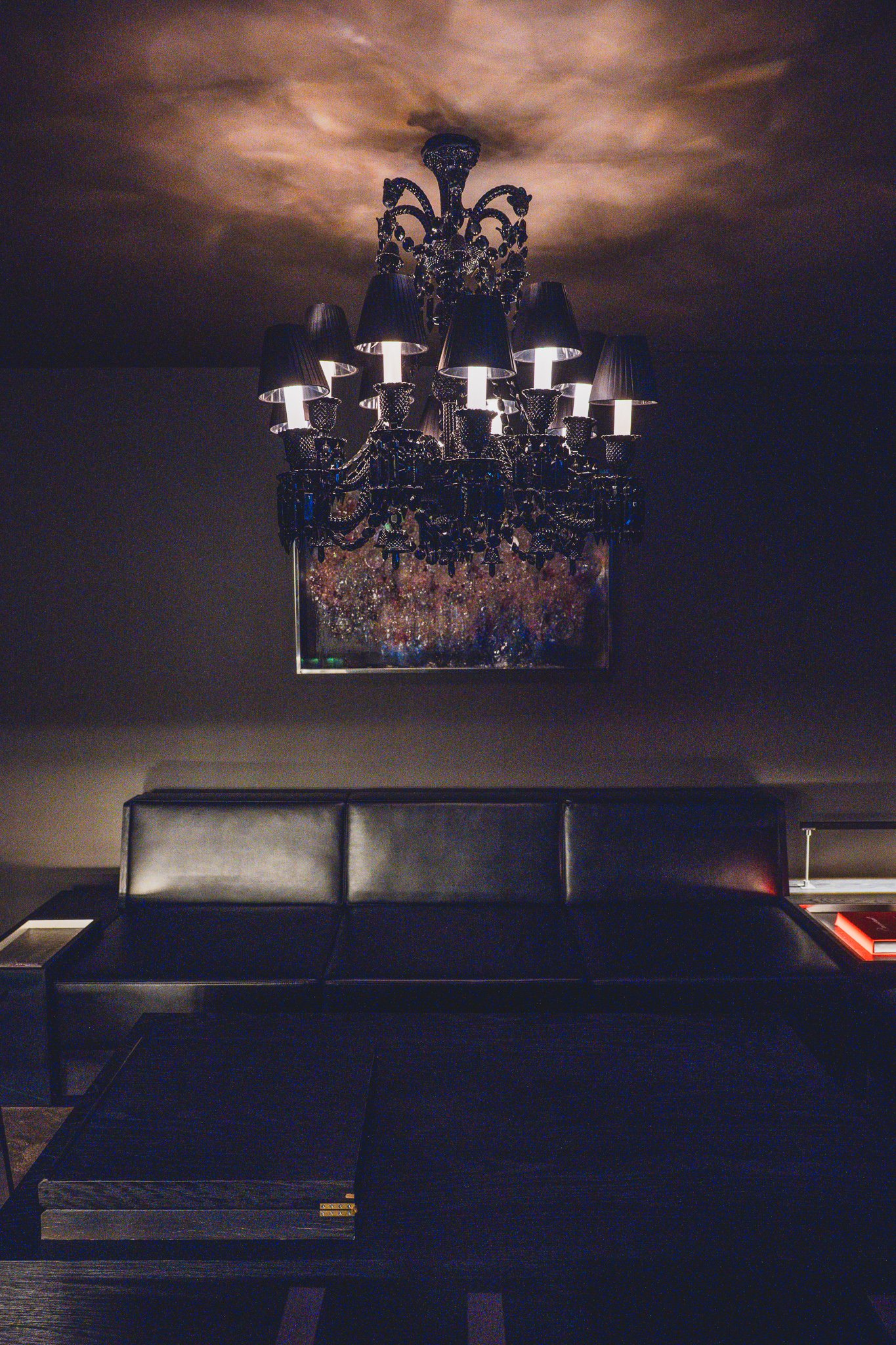 A dark, moody lounge room with a black crystal chandelier over leather sofas and an artwork on the wall