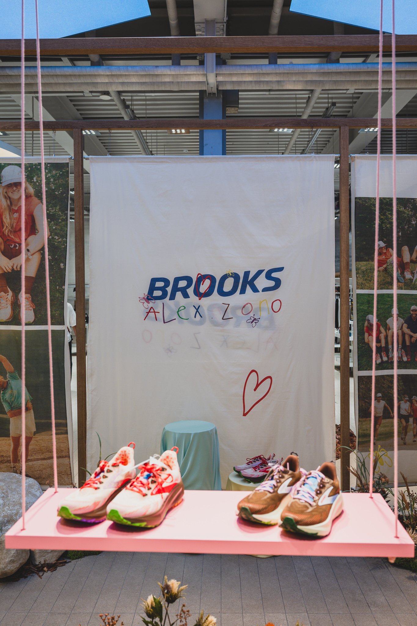 Fabric backdrop with the Brooks Alex Zono logo, two pairs of Ghost Trail shoes displayed on a pink swing shelf