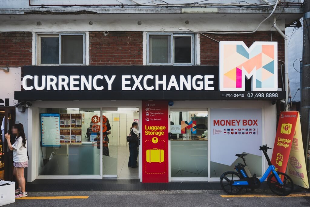 The storefront of Money Box Seongsu Station branch in Seoul, a black sign reading "CURRENCY EXCHANGE" above a glass shopfront with an electronic rate board in the window and a red pillar advertising luggage storage