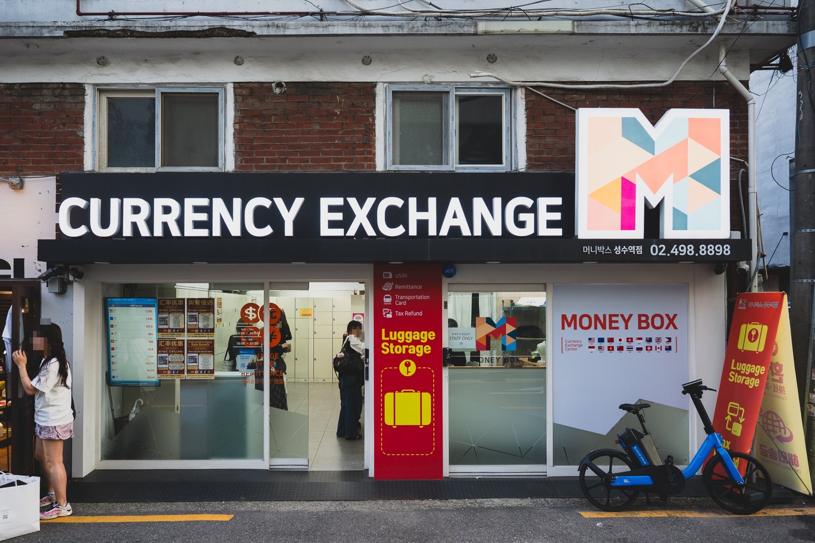 The storefront of Money Box Seongsu Station branch in Seoul, a black sign reading "CURRENCY EXCHANGE" above a glass shopfront with an electronic rate board in the window and a red pillar advertising luggage storage