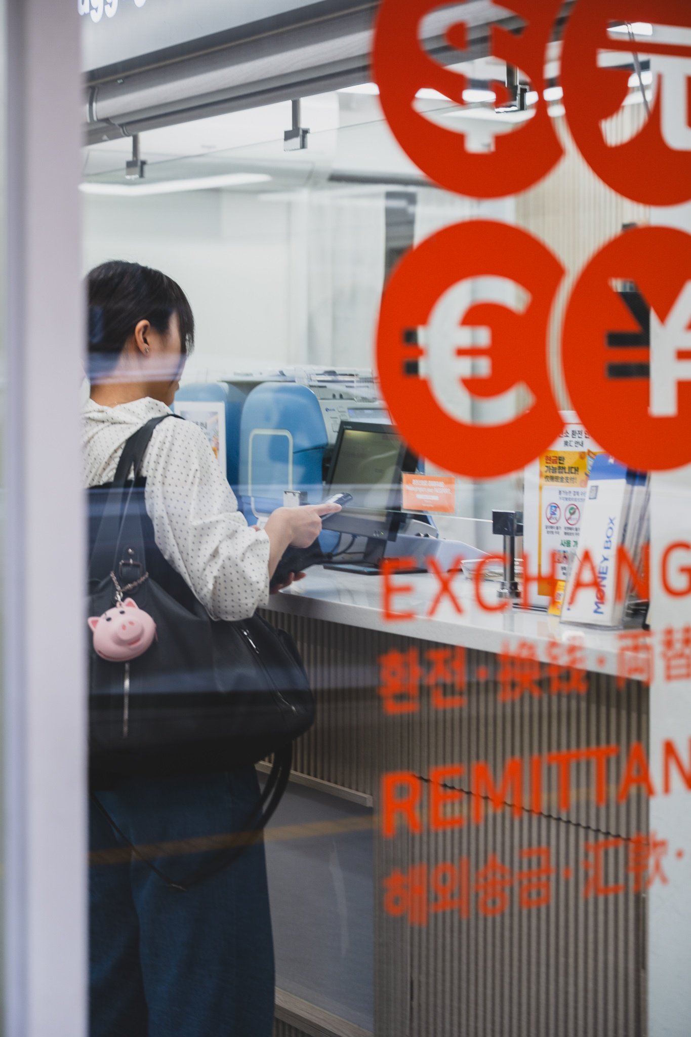 View through the shop window of the exchange counter, a customer standing at the service window with dollar, euro and yen symbols decaled on the glass alongside the words EXCHANGE and REMITTANCE
