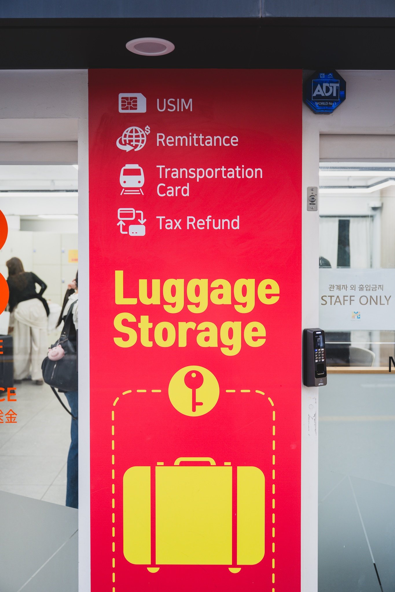 A red pillar sign at the shop entrance listing USIM, Remittance, Transportation Card and Tax Refund services, with "Luggage Storage" in large yellow lettering below