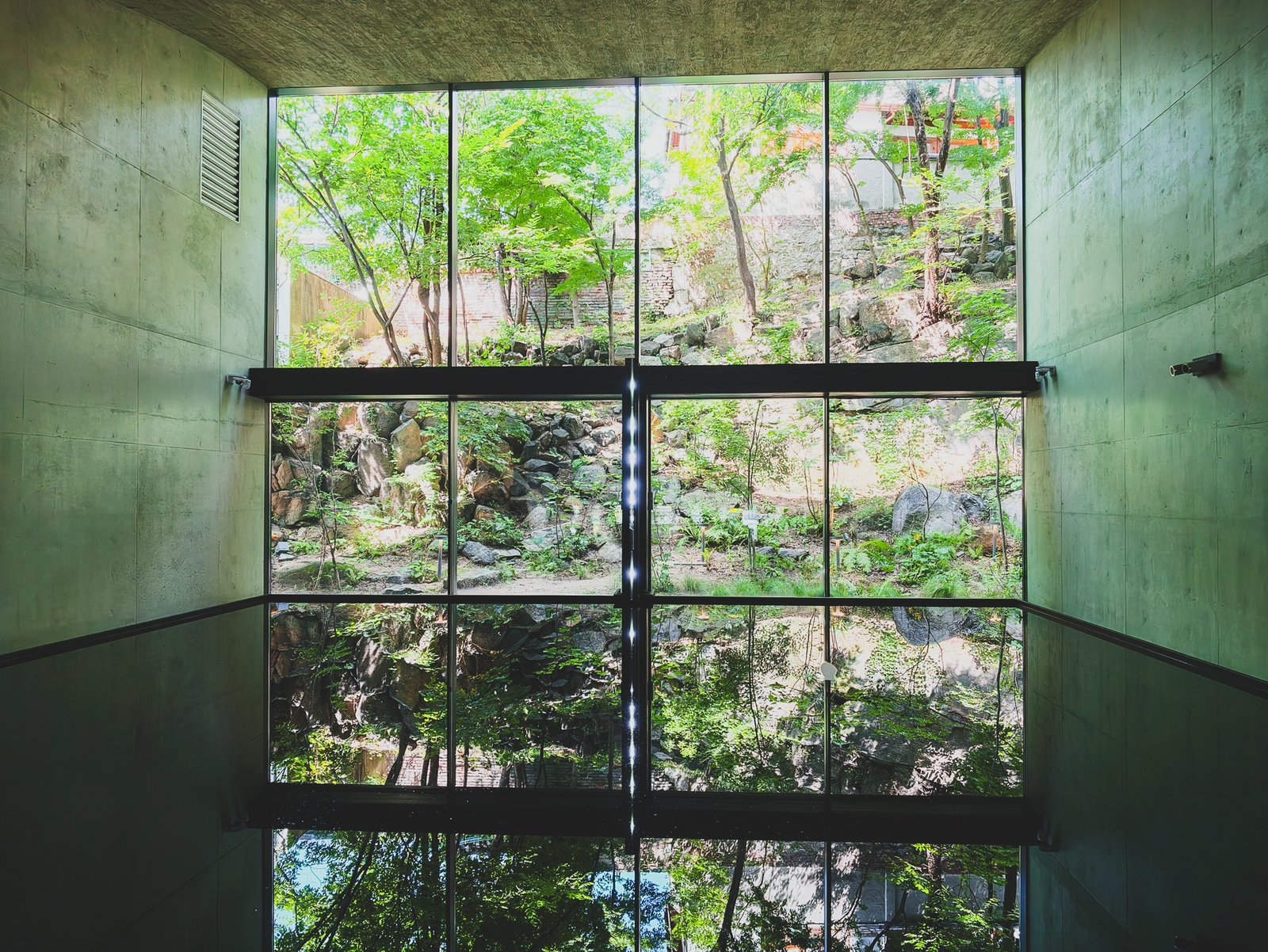 Floor-to-ceiling glass wall overlooking Bukchon's garden and trees, a single strobe of light reflected in a shallow floor pool