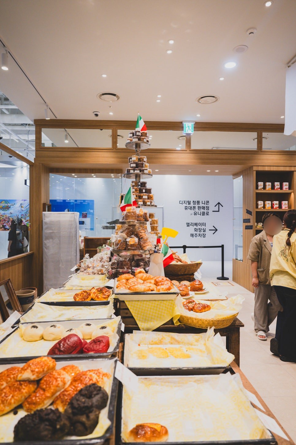 A long bread display counter stacked with croissants, savory pastries, and a tiered tower of packaged cookies topped with a small Italian flag