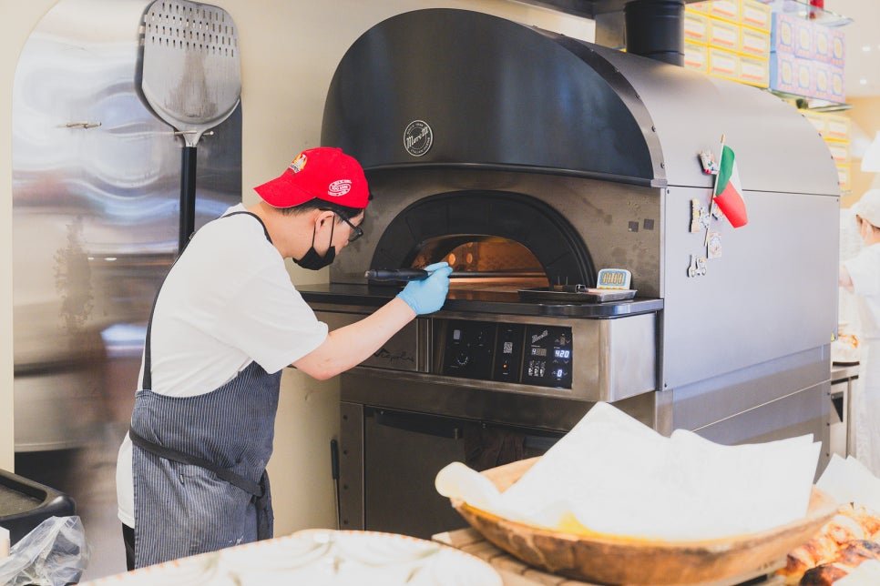 A chef sliding dough into a large wood-fired oven, with an Italian flag magnet stuck to the oven door