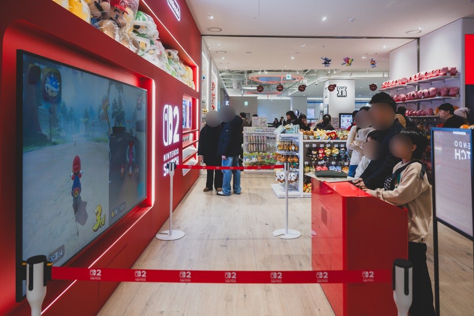 Kids and adults playing Mario Kart on a large demo screen at a Switch 2 play station, roped off with red barriers