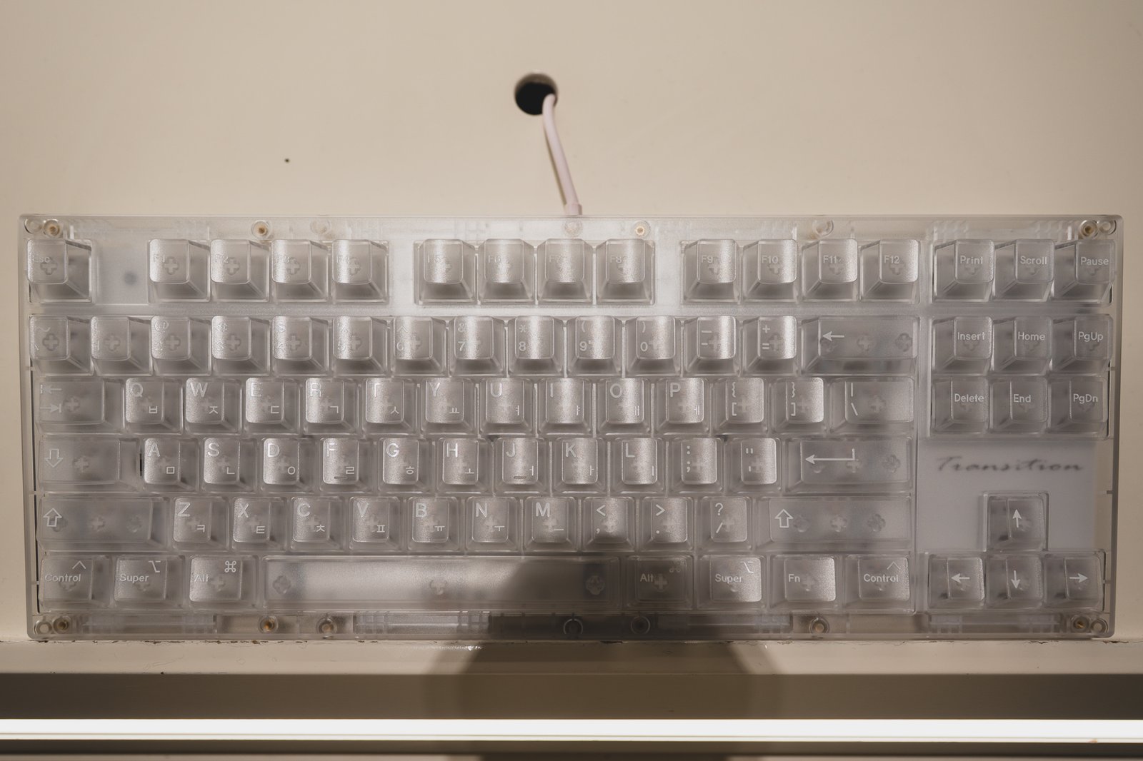 Close-up of a translucent white mechanical keyboard with a "Transition" nameplate, backlit against a beige wall