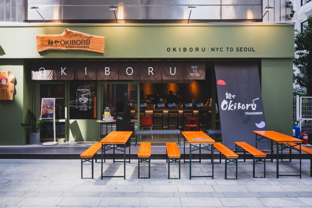 OKIBORU's olive-green storefront with a wooden "麺や OKIBORU" sign and outdoor bench seating